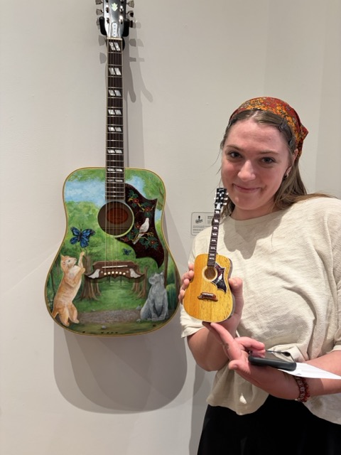 Dogwood Arts Youth Arts Program Artist Holds Her Mini Guitar by AXE HEAVEN® Next to the Full-Size Acoustic Guitar She Painted in 2026: Where the Forest Plays